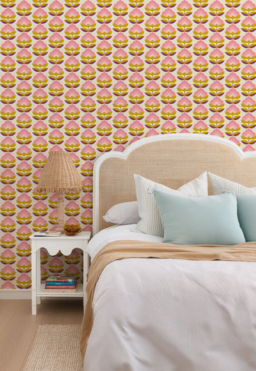 Retro Lotus Wallpaper featuring a repeating pattern of stylized lotus flowers in shades of pink, golden yellow, and deep purple on a cream background, installed on a bedroom wall. The wallpaper is visible behind a bed with a woven rattan headboard, white and light blue pillows, and a striped duvet. A wicker lamp and a white nightstand with books are also visible.