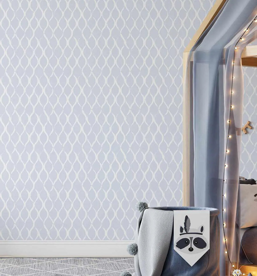 "Ripples wallpaper featuring a repeating pattern of hand-painted, elongated ovals in off-white on a soft periwinkle blue background, creating a subtle wave-like texture. The wallpaper is displayed in a child's bedroom with a light wood canopy bed draped with sheer blue fabric and string lights. A grey storage basket with a raccoon face design sits in the foreground."