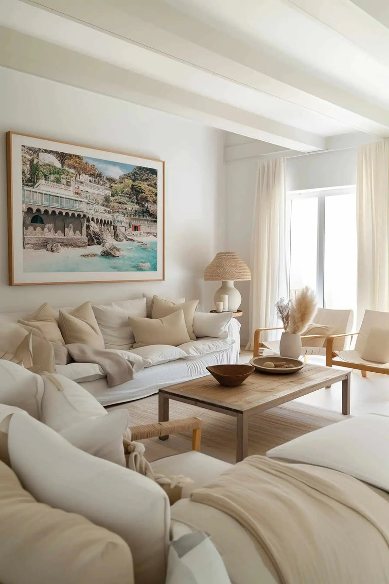 Large framed art print displayed above a white slipcover sofa in a bright, minimalist living room. The print is a photograph of a coastal Italian scene with multi-story white buildings built into a rocky hillside, with turquoise water and a sandy beach in the foreground, and a natural wood frame with a white border. The room features cream-colored curtains, a woven lamp, and two light wood armchairs.