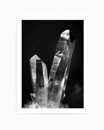 Black and white photograph of three clear quartz crystals against a dark background. The crystals are tall and pointed, with visible facets and internal inclusions, reflecting light to create bright highlights and deep shadows. The composition emphasizes the natural texture and sharp forms of the minerals.