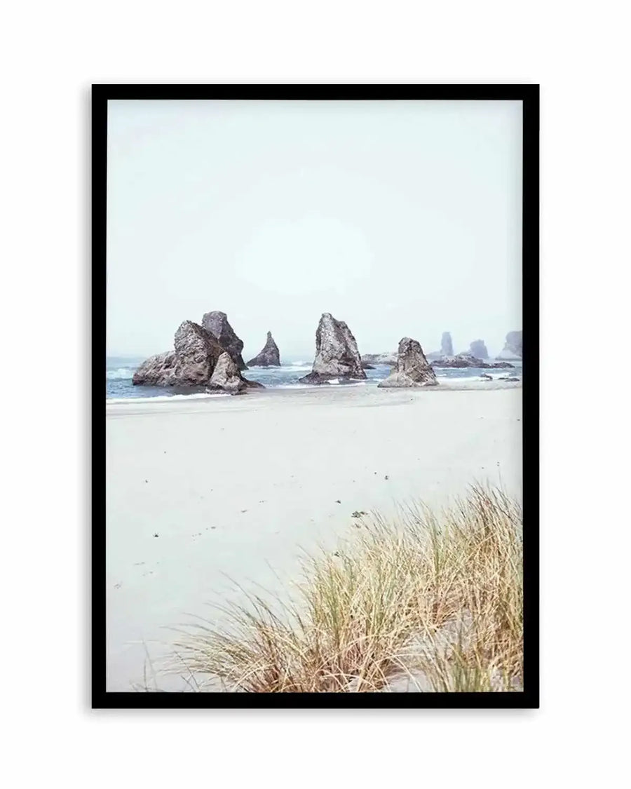 Framed photography art print of a serene coastal beach scene with a black frame and white border. The composition features several large, dark grey rock formations emerging from the light blue ocean, with gentle waves breaking on a wide expanse of pale sandy beach. In the foreground, clumps of dry, light brown and green beach grass grow in the sand under an overcast, light grey sky.