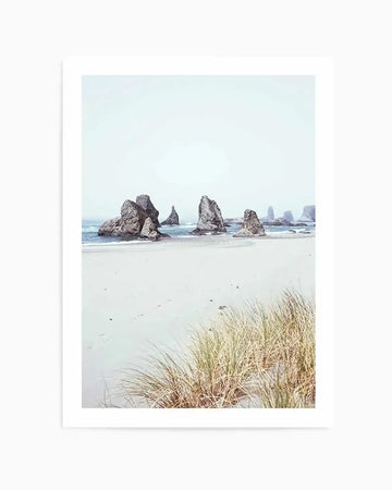 Framed photography art print of a serene coastal beach scene with a natural wood frame and white border. The composition features several large, dark grey rock formations emerging from the light blue ocean and wet, pale sand under a soft, overcast sky. In the foreground, clumps of dry, golden-brown beach grass grow on the sand.