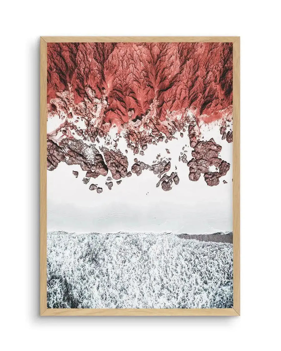Framed aerial photography art print of a rocky coastline with a natural oak frame and white border. The composition features a vibrant red-orange rocky cliff face meeting a white sandy beach, with foamy white ocean waves crashing onto the shore.