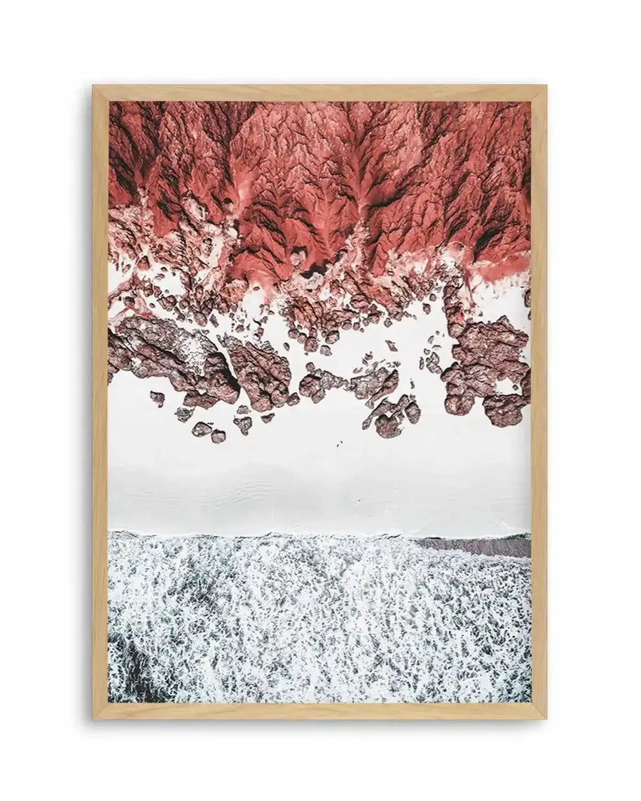 Aerial photography art print of a rocky coastline with a natural oak frame and white border. The image captures the vibrant reddish-brown and terracotta tones of the rugged, textured cliffs and scattered rocks meeting the bright white foamy waves of the ocean. The composition shows the land on the upper half, a thin strip of white sand, and the ocean with breaking waves on the lower half.