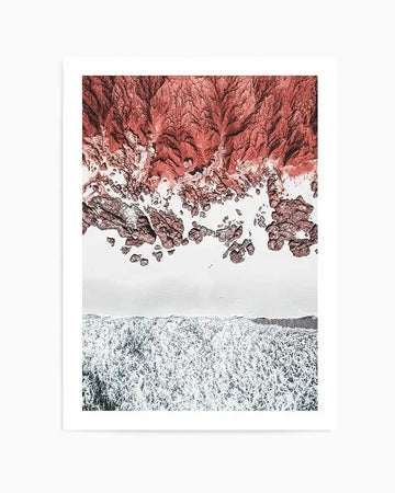 Aerial photography art print of a rocky coastline with a wide white beach and crashing waves, printed on fine art paper with a white border. The top third features reddish-brown, textured rock formations resembling a rugged, eroded landscape. Below this, a smooth, light grey sandy beach separates the rocks from the ocean. The bottom third shows the ocean with white foamy waves breaking onto the shore, creating a dynamic contrast between the land and sea.