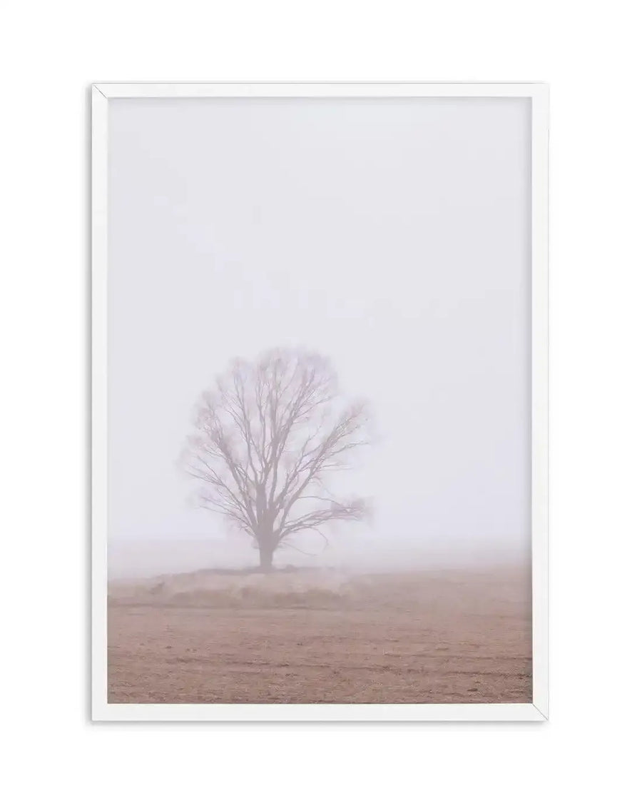 Framed photography art print of a solitary tree in a misty, muted landscape, with a white frame and white border. The bare tree stands in the center, its branches reaching upwards, while the ground below is a soft, earthy brown, fading into the pervasive light grey mist that fills the background.