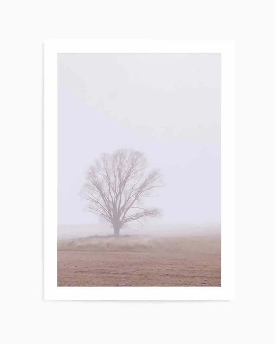 Framed photography print of a single, bare tree in a field on a foggy day, with a white border. The tree, with its dark, intricate branches, stands in the middle ground, partially obscured by a thick, pale lavender mist that fills the upper two-thirds of the frame. The foreground features a flat, reddish-brown field with subtle texture.
