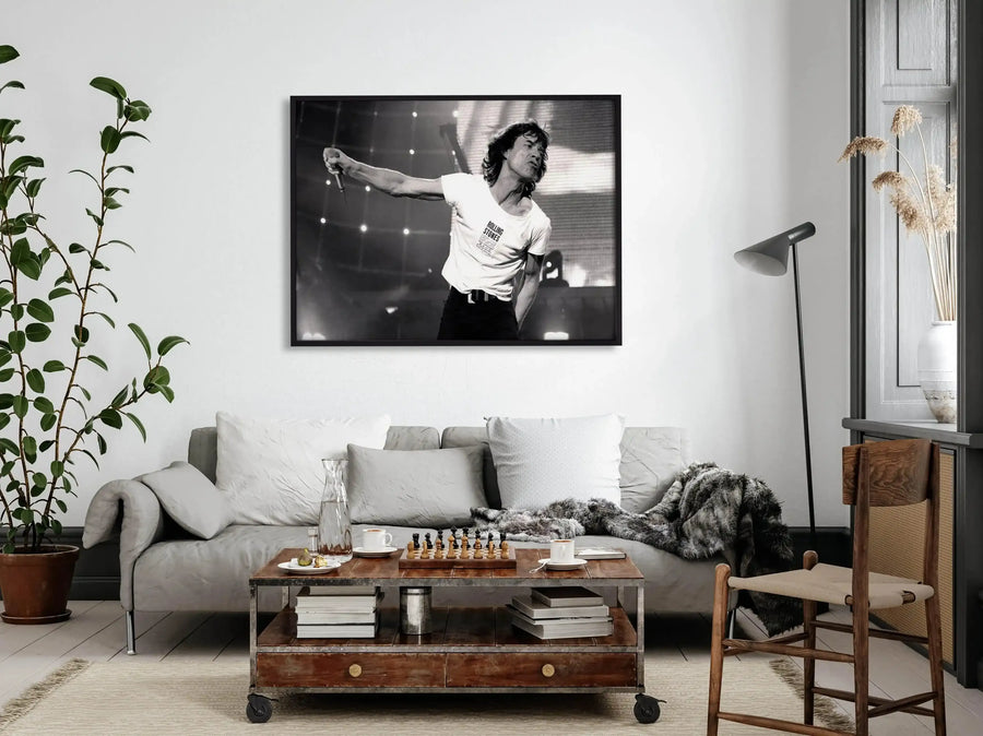 Lifestyle shot of a framed black and white photograph of Mick Jagger performing live, displayed above a grey sofa in a modern living room. The print features Mick Jagger in a white t-shirt with "ROLLING STONES" visible, holding a microphone and looking to the left with a dynamic expression. The room includes a potted plant, a wooden coffee table with a chess set, books, and a glass of water, and a dark grey floor lamp.