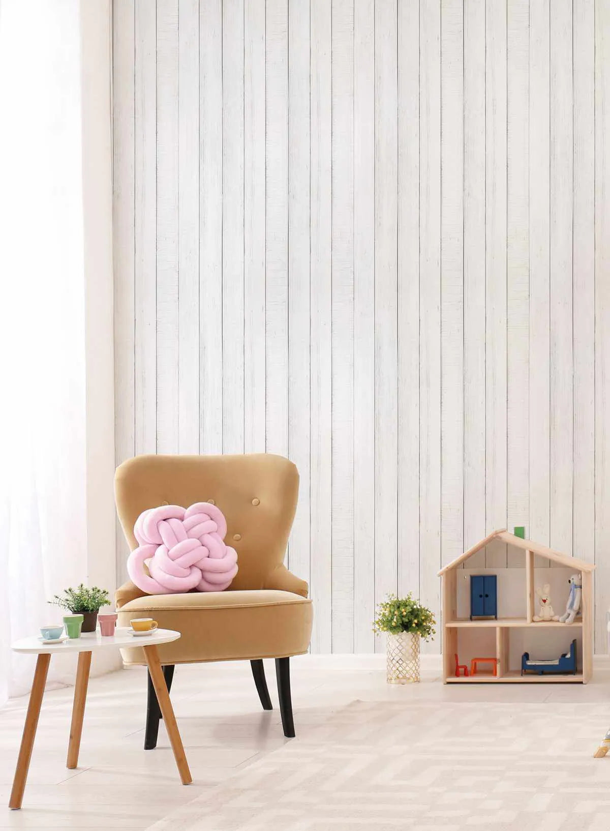 Lifestyle shot of a child's room featuring a white wood panel wallpaper. The wallpaper has vertical planks with subtle grey lines indicating the joins and a slightly distressed, rustic texture. The room includes a mustard yellow armchair with a pink knot cushion, a small white side table with miniature teacups, and a wooden dollhouse with blue doors and miniature furniture.