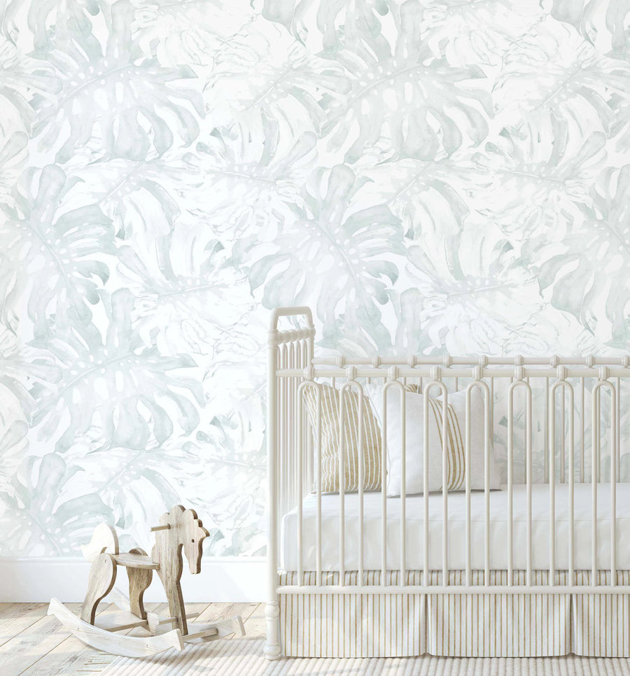 Lifestyle shot of a nursery featuring Sage Monstera Wallpaper. The wallpaper has a repeating pattern of large, hand-painted monstera leaves in a muted sage green and pale grey on a soft white background, with a loose, organic watercolor quality. A white metal crib with vertical bars and a striped beige and white skirt is positioned against the wallpaper, with a wooden rocking horse on the light wood floor in the foreground.