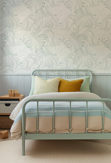 Lifestyle shot of a child's bedroom featuring a wallpaper with a repeating pattern of large-scale monstera leaves in a muted sage green and pale blue against a soft white background. The wallpaper has a watercolour quality with visible brushstroke texture. The wallpaper is displayed above a light blue wainscoting and a light green metal bed frame with a striped duvet in shades of blue, cream, and beige. A wooden bedside table with building blocks sits to the left of the bed.