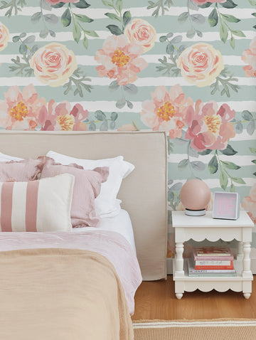 Peel and stick wallpaper featuring a repeating pattern of horizontal sage green stripes with large-scale watercolour flowers in shades of blush pink, peach, and cream, with green leaves and branches. The wallpaper is displayed behind a bed with a beige upholstered headboard, striped pink and white bedding, and a white nightstand with a pink spherical lamp and a light box.