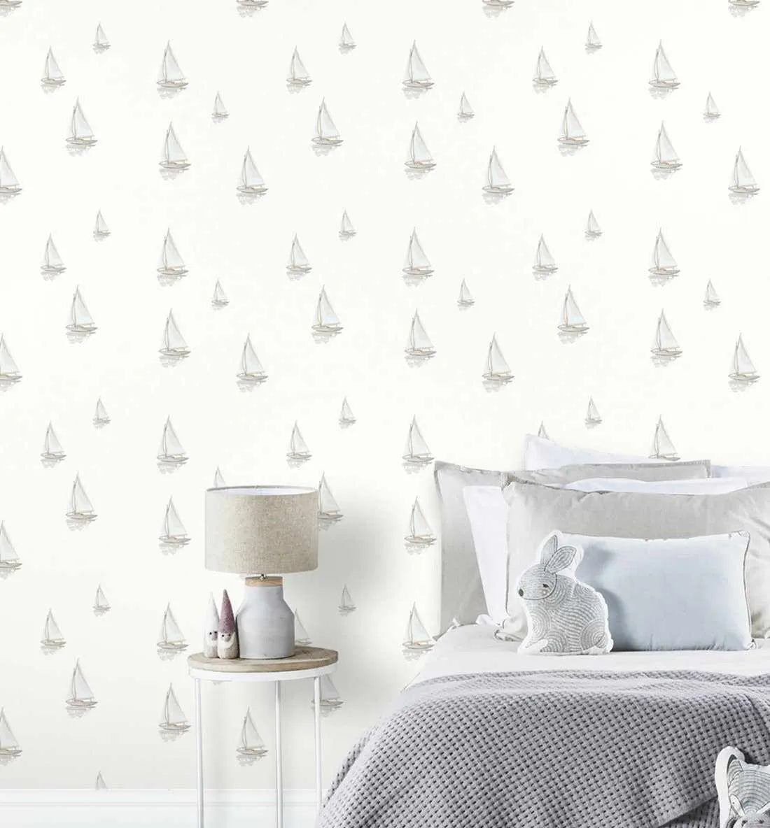 Lifestyle shot of a bedroom featuring peel and stick wallpaper with a repeating pattern of hand-painted sailing boats in muted grey and white with subtle reflections on a soft white background. The wallpaper is displayed behind a bed with grey and white bedding, a grey knitted throw, and a white rabbit-shaped cushion. A bedside table with a lamp and small decorative items is visible to the left.