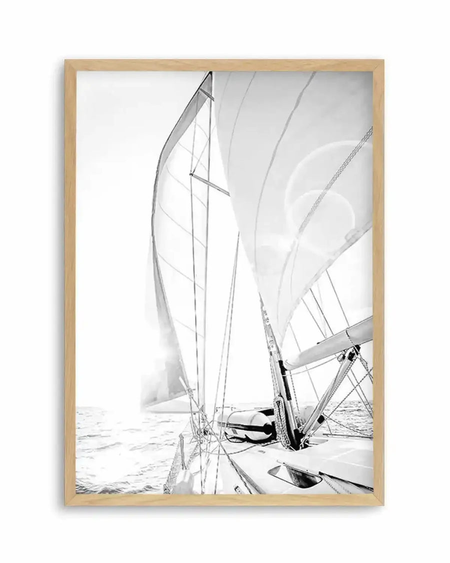 Framed black and white photograph of a sailboat on the ocean with natural wood frame and white border. The composition features the bow of the sailboat with its sails unfurled, catching the bright sunlight, creating lens flare and reflections on the water.