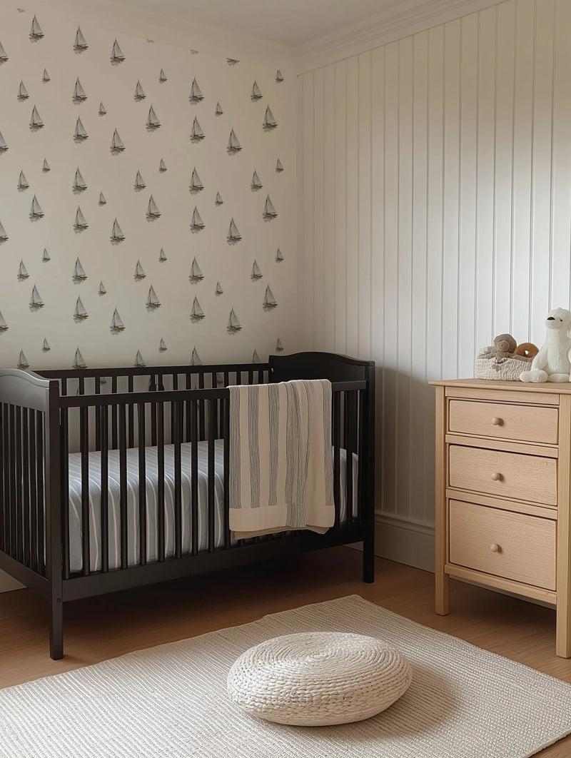 Lifestyle shot of a nursery with a dark wood crib against a white wall featuring sailing boat wallpaper. The wallpaper has a repeating pattern of hand-drawn sailboats with white sails and dark hulls, appearing to float on calm water with subtle reflections. The room includes a light wood dresser with three drawers, a white woven rug, and a round white knitted pouf on a light wood floor. A striped blanket in cream and black hangs over the crib.