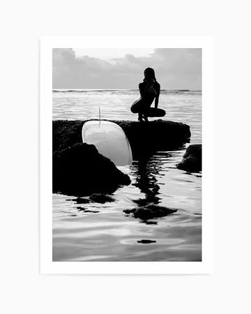 Black and white photographic art print titled "Salty II" by Mario Stefanelli, featuring a silhouetted female surfer in a wetsuit crouching on dark rocky outcrops along a misty beach. She holds a white surfboard with a visible fin. Moody seascape with calm grey waters and overcast sky in the background. High contrast monochromatic imagery with dramatic lighting. Displayed in a white frame with white border.