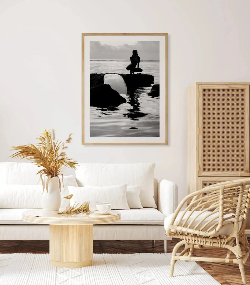 Black and white photograph titled "Salty II" by Mario Stefanelli, framed in natural wood with white border. The image shows a surfer in a wetsuit crouching on dark rocks in shallow ocean water, holding a white surfboard. Misty coastline and calm water visible in the background. Displayed on white wall above a cream linen sofa with natural wood rattan chair and small cylindrical side table holding dried pampas grass in white ceramic vase.