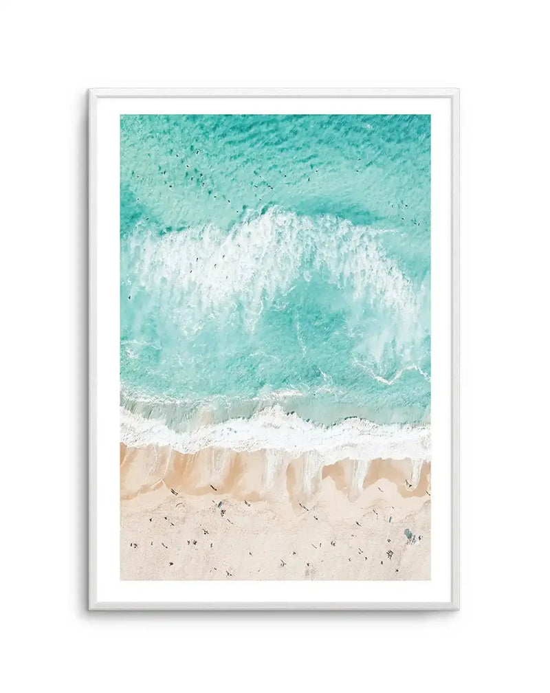 Framed aerial photograph of a beach scene with a white frame and white border. The composition features the turquoise ocean with white waves crashing onto a sandy beach, with tiny figures of people scattered along the shore.