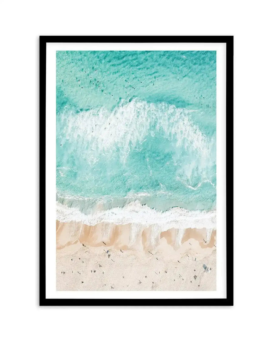 Framed aerial photograph of Bondi Beach with a black frame and white border. The composition shows the turquoise ocean with white waves breaking on a light beige sandy beach, with tiny dark figures of people scattered across the sand and in the water.