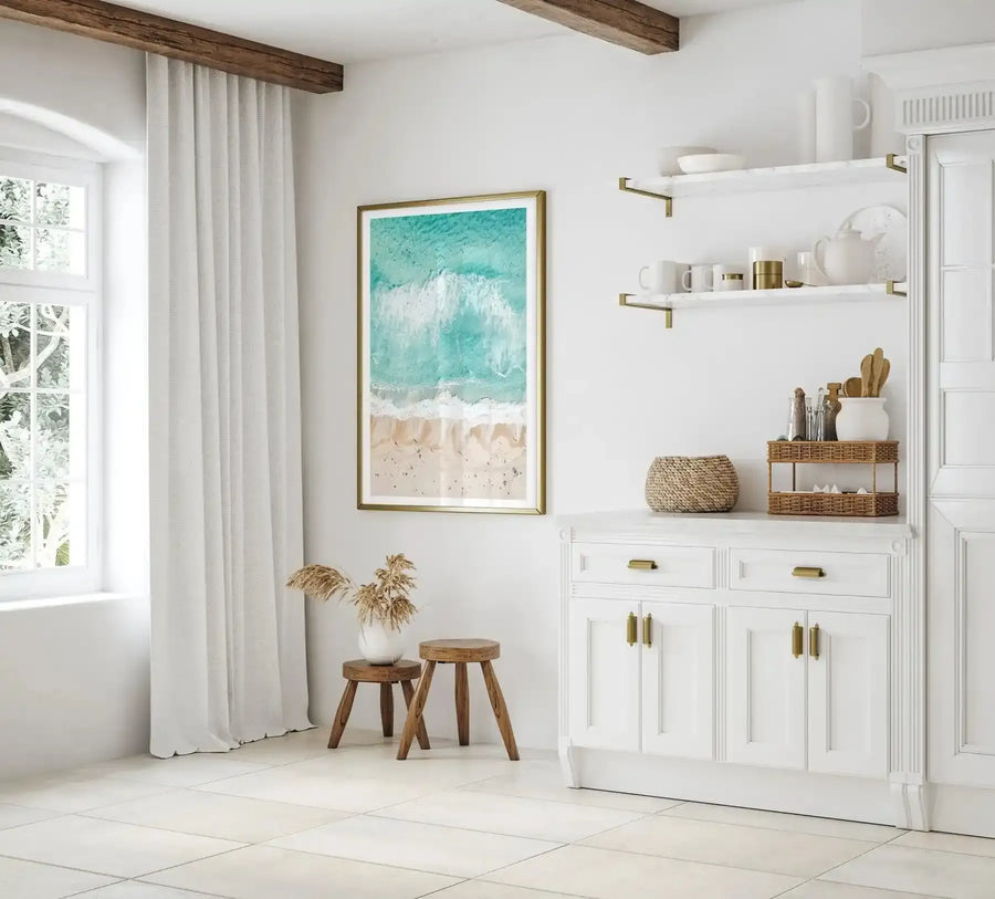 Aerial photograph art print titled Sand to Sea Bondi displayed in a gold frame with white border, hung on a white wall above wooden stools. The print shows a turquoise ocean with white wave foam and sandy beach dotted with swimmers, captured from above. The room features white cabinetry, floating shelves, cream curtains, exposed wooden ceiling beams, and woven basket accessories, creating a coastal aesthetic.