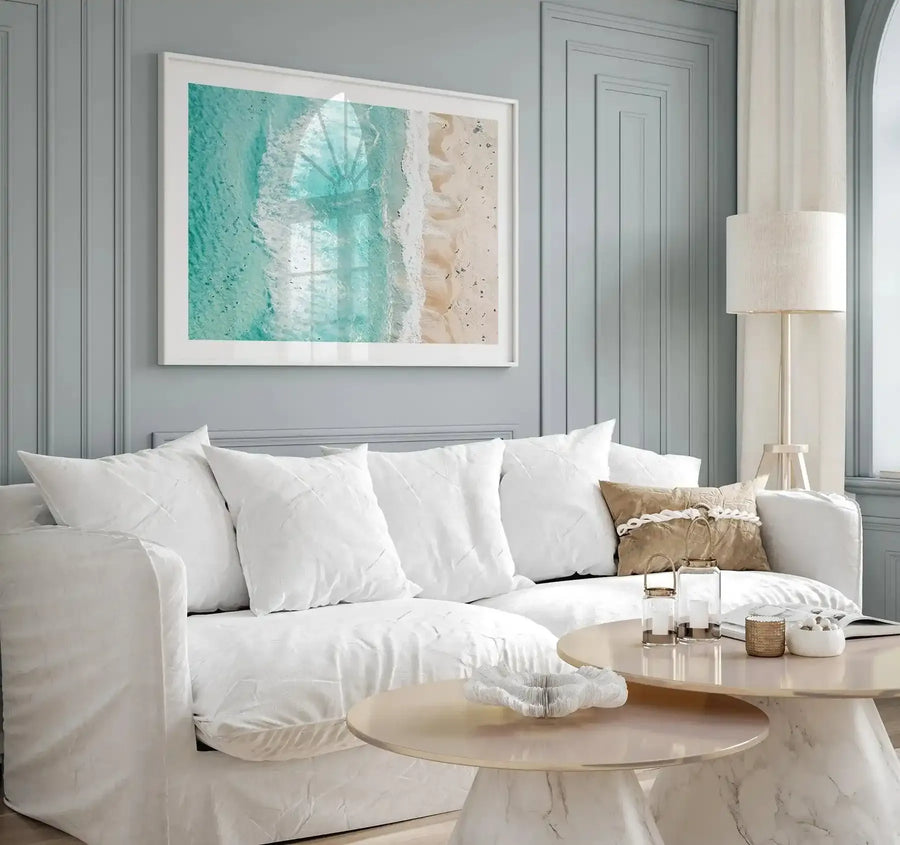 Aerial photograph framed in white showing turquoise ocean water, white foam waves, and sandy beach in warm beige tones. The image captures a coastal landscape from above with a soft, serene quality. White wooden frame with white border displayed above a cream linen sofa styled with white pillows and natural wood accent table.