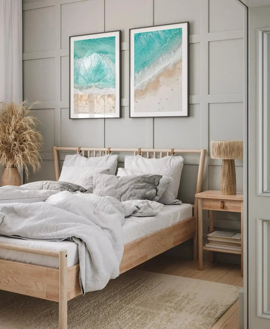 Two framed aerial photography art prints displayed side-by-side above a wooden bed in a neutral-toned bedroom. The prints feature a bird's-eye view of a beach with turquoise ocean waves crashing onto light beige sand, dotted with tiny figures of people. Both prints have a black frame and white border. The room includes a light wood bed frame with white and striped linen bedding, a large vase of dried pampas grass, a wooden bedside table with a woven lamp, and a textured beige rug.
