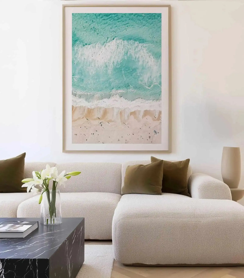 Lifestyle shot of a minimalist living room featuring a large framed aerial photo wall art print displayed above a textured cream sectional sofa. The print, titled "Sand to Sea Bondi," captures a vibrant turquoise ocean with white crashing waves meeting a sandy beach dotted with tiny figures, framed in natural wood with a white border. The room includes dark olive green throw pillows and a black marble coffee table with a vase of white lilies.