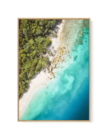 Aerial view framed canvas print showing a tropical coastline where dense green forest meets a pristine white sand beach and turquoise ocean. Large golden boulders dot the shoreline. The water transitions from pale aqua near the shore to deeper teal and blue in deeper areas. Natural wood frame with no border. Photograph-style imagery capturing a coastal landscape from above.