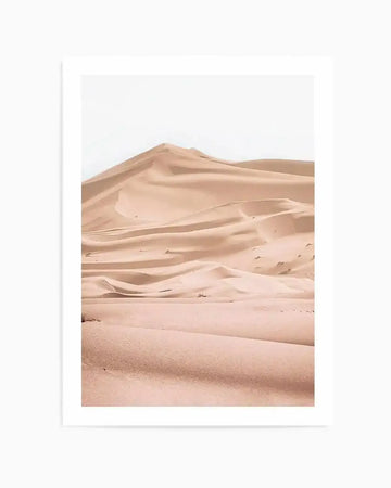 Framed photography art print of a desert landscape with rolling sand dunes in shades of pale peach, dusty rose, and warm beige under a bright white sky. The composition features soft, undulating curves and subtle shadows highlighting the natural texture of the sand.