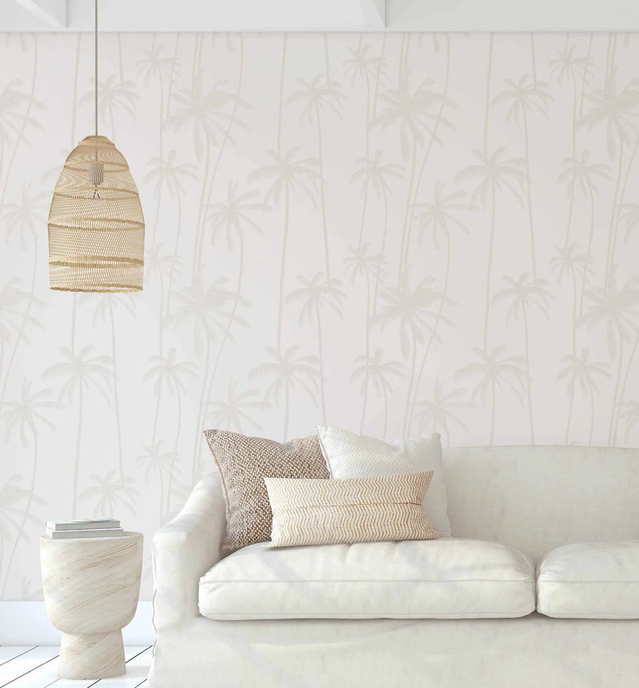Lifestyle shot of a minimalist living room featuring a white sofa with neutral-toned cushions and a light wood side table, set against a wall covered in Sandy Palms Wallpaper. The wallpaper features a repeating pattern of tall, slender palm trees with delicate fronds in a subtle, light beige hue on a soft white background, creating a serene, tropical ambiance. A woven pendant light hangs in the foreground.