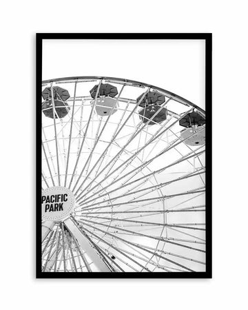 Black and white art print depicting a towering Ferris wheel photographed from below, showing the iconic "Pacific Park" text on the hub. The composition captures the geometric steel framework and support cables radiating outward, with four gondola cars visible at the top against a bright sky. The image has high contrast with crisp details of the mechanical structure. The print is mounted in a black wooden frame with a white border.