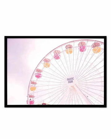Framed art print featuring a close-up photograph of the Pacific Park Ferris wheel in Santa Monica. The iconic structure displays passenger gondolas in soft shades of blush pink, coral, and pale yellow against a white sky. The composition captures the geometric spoke pattern radiating from the center hub, with "PACIFIC PARK" text visible. The photograph has a dreamy, pastel aesthetic. The print is displayed in a black frame with white border.
