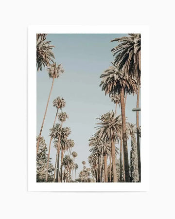 Photography art print featuring a low-angle view of tall palm trees lining a street against a clear, pale blue sky. The composition shows two rows of palm trees, with the fronds of the trees on the right appearing closer and more detailed, while those on the left recede into the distance. The overall mood is bright and airy, with a vintage, slightly desaturated colour palette.