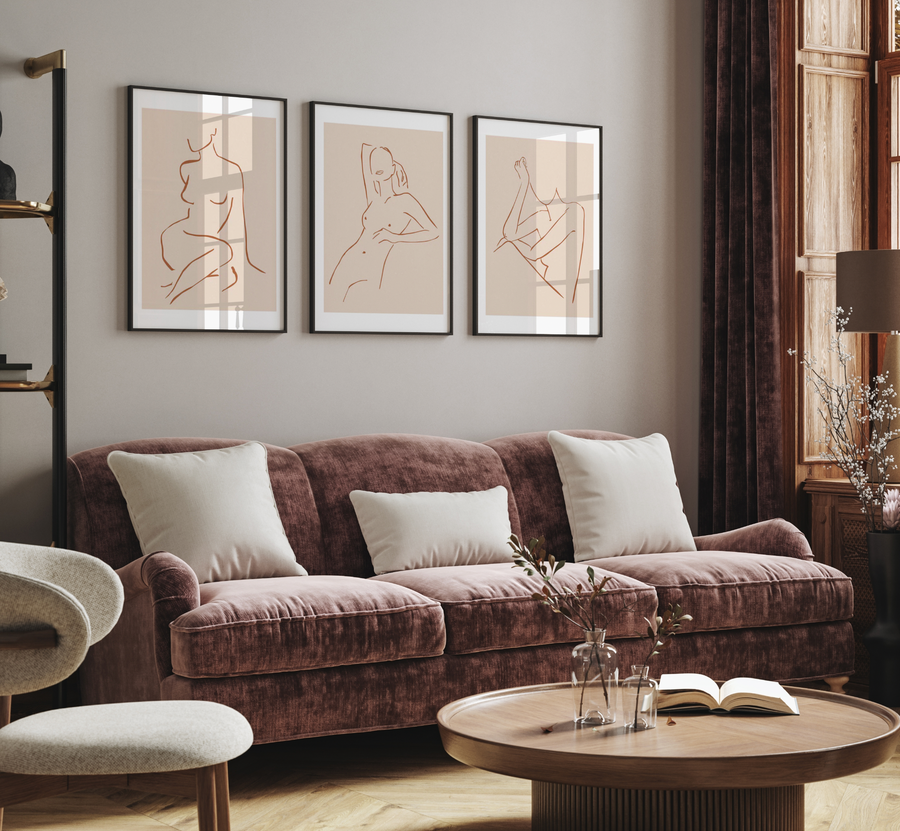 Lifestyle shot of a living room with three framed art prints displayed above a velvet sofa. The prints feature minimalist line art illustrations of female forms in terracotta on a warm beige background, with black frames and white borders. The room includes a dusty rose velvet sofa with cream cushions, a round wooden coffee table with a book and vases, and dark brown curtains next to a wooden window.