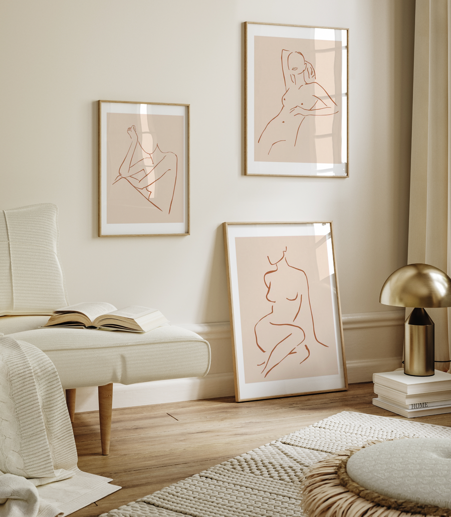 Lifestyle shot of three framed art prints in a minimalist room with a cream armchair and gold lamp. The prints feature abstract line drawings of female forms in terracotta on a light peach background, with natural wood frames and white borders. The largest print, leaning against the wall, shows a seated figure. The two smaller prints, hung on the wall, depict a seated figure with arms raised and a figure with one arm raised. The room has light cream walls, a light wood floor, and a cream textured rug.