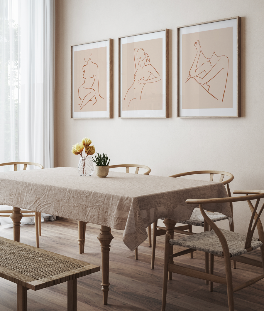Lifestyle shot of a dining room featuring three framed art prints displayed above a wooden dining table with a linen tablecloth. The art prints feature minimalist line drawings of female forms in terracotta brown against a warm cream background, with natural wood frames and white borders. The room includes wooden dining chairs with woven seats, a wooden bench, and a vase of dried yellow flowers and a potted plant on the table.