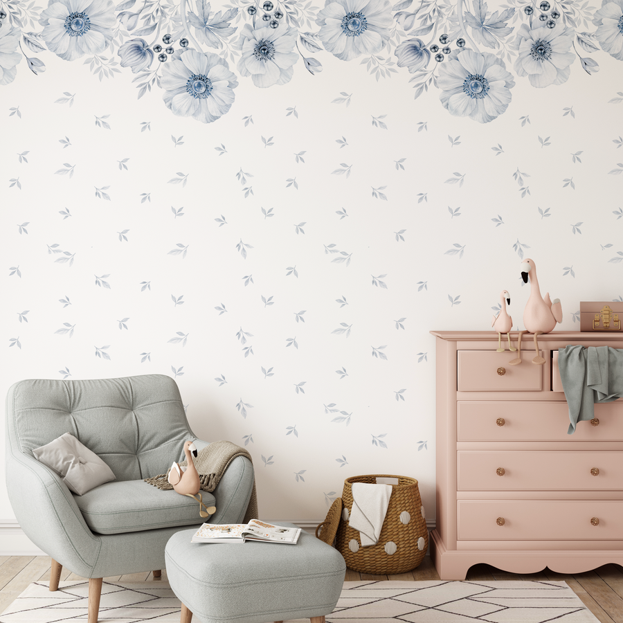 Nursery:
Peel and stick wallpaper featuring a repeating pattern of large-scale blue anemone flowers with dark blue centers and delicate grey-blue leaves, arranged in a border along the top of the wall, with smaller individual leaves scattered across the rest of the soft white background. The room includes a light grey armchair and ottoman, a pink dresser with gold knobs, a woven basket, and two pink flamingo plush toys.