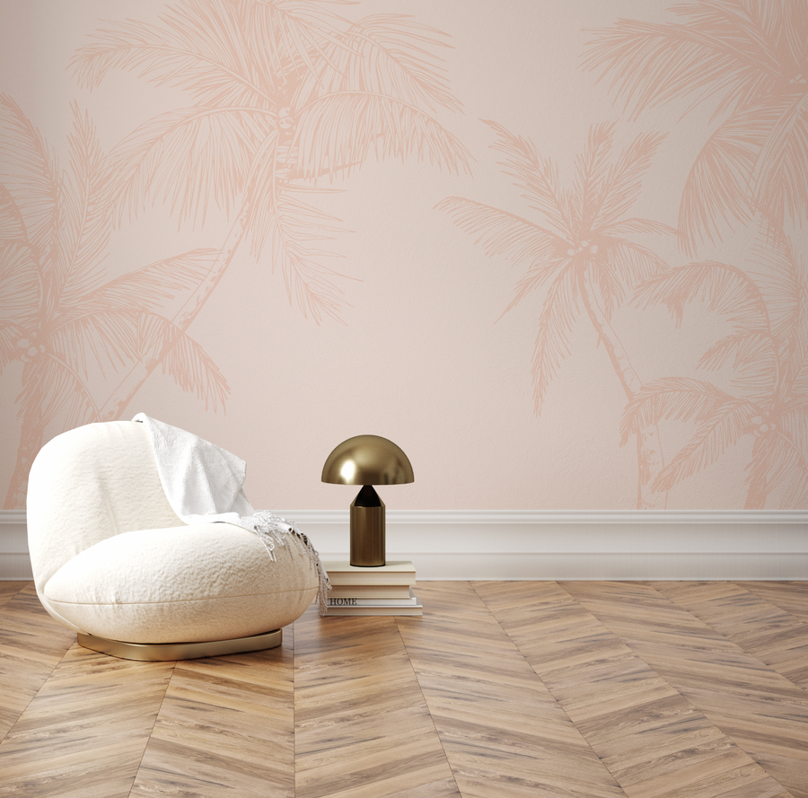 Lifestyle shot of a minimalist room featuring a wallpaper mural with a repeating pattern of hand-drawn palm trees in a soft terracotta pink on a pale blush pink background. The wallpaper has a subtle textured finish. A cream-colored bouclé armchair with a white throw blanket is positioned in the foreground, next to a stack of books and a brass mushroom-shaped lamp. The room has light wood flooring in a herringbone pattern and white baseboards.
