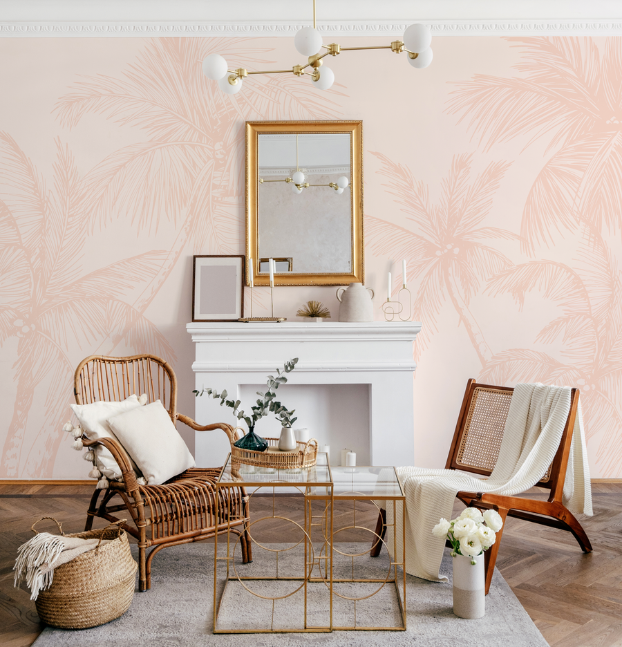 Lifestyle shot of a living room featuring a wallpaper mural with a repeating pattern of hand-drawn palm trees in a soft terracotta pink on a light blush background. The room includes a white fireplace, a gold-framed mirror, a rattan armchair with cream cushions, a wooden chair with cane webbing, and gold accent tables.