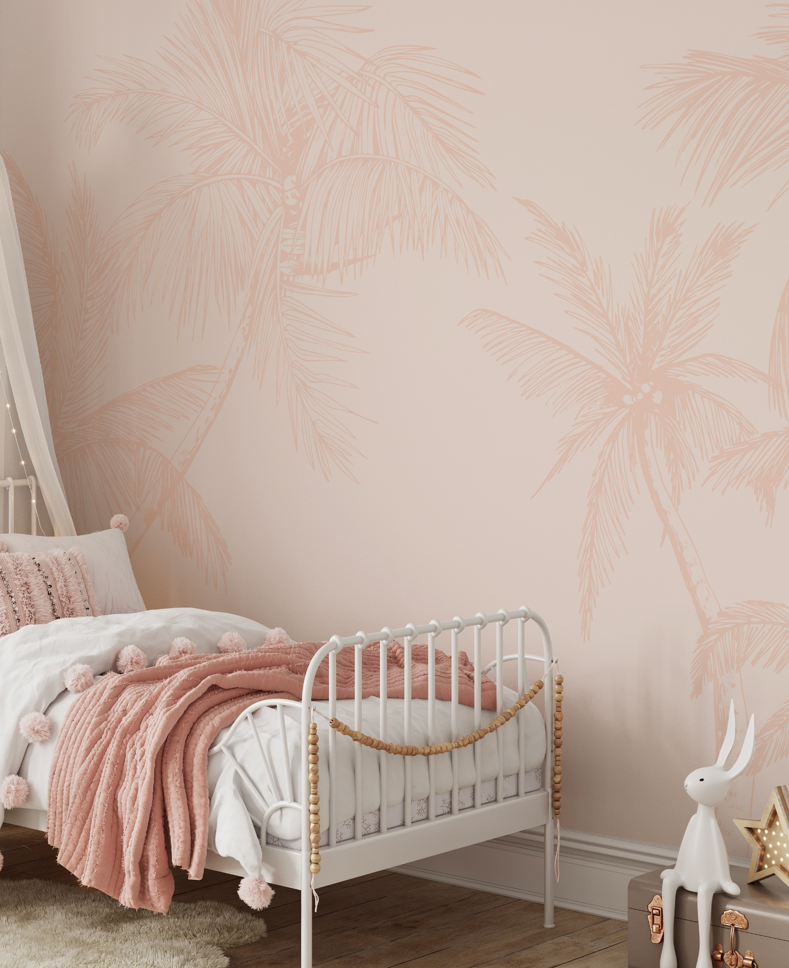 Lifestyle shot of a child's bedroom featuring a wallpaper mural with large-scale, hand-drawn palm trees in a soft terracotta pink on a lighter, muted pink background. The wallpaper covers the entire visible wall behind a white metal toddler bed with a blush pink quilted blanket, white bedding, and a white canopy with fairy lights. A white rabbit figurine sits on a grey trunk next to a star-shaped light on the wooden floor.