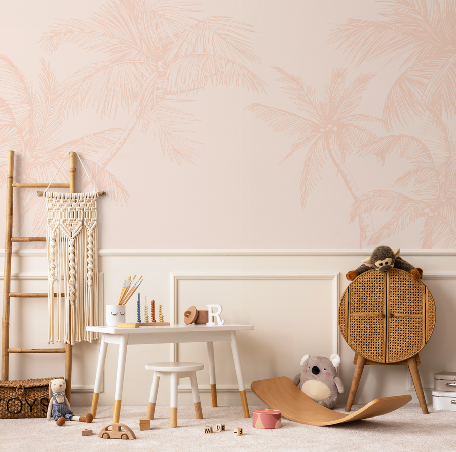 Lifestyle shot of a child's playroom featuring a wallpaper mural with a repeating pattern of hand-drawn palm trees in a soft terracotta pink on a pale pink background. The room includes a small white table and stool, a woven storage basket, a macrame wall hanging, and various wooden toys.