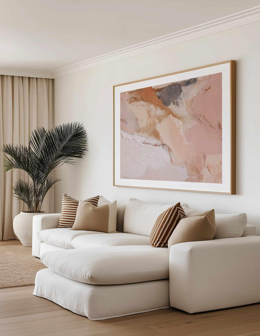Sculpted Clay I art print displayed in a natural wood frame with white border, hung on a cream wall above a curved white upholstered sofa. The artwork features abstract brushstrokes in dusty rose, warm terracotta, soft cream, and charcoal gray tones, creating an organic, textured composition with visible brushwork and layered paint effects. The room styling includes striped throw pillows in tan and brown, a potted black palm plant, beige curtains, and light wood flooring, creating a warm, neutral aesthetic.