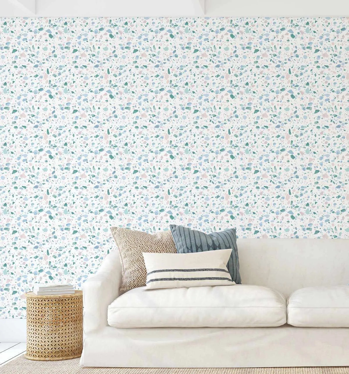 Sea Glass Terrazzo Wallpaper displayed on a living room wall behind a cream-colored sofa. The wallpaper features a dense, scattered terrazzo pattern with small irregular stone-like shapes in shades of sage green, dusty blue, soft pink, and gray on a white background. The design has a speckled, granite-like appearance with a matte finish. Three decorative pillows in neutral tones sit on the sofa, and a woven cylindrical side table is positioned to the left.