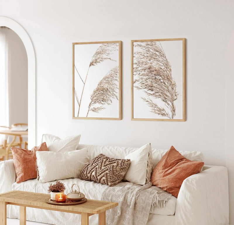 Lifestyle shot of two framed art prints displayed above a white sofa in a bright, minimalist living room. Each print features a close-up photograph of dried pampas grass in a natural, light brown hue against a clean white background, with natural wood frames and white borders. The sofa is adorned with cream and rust-coloured cushions, a textured brown cushion, and a light-coloured throw blanket. A woven wooden bench in front of the sofa holds a lit candle, a small plant, and a decorative teapot.