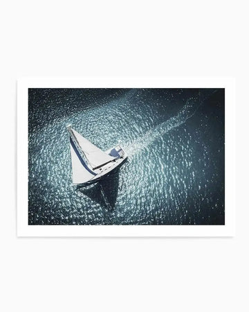 "Art print featuring an aerial photograph of a white sailboat with its sail up, cutting through dark blue water under bright sunlight, creating a shimmering wake and a long shadow on the water. The composition captures the dynamic movement of the boat and the reflective quality of the ocean surface."