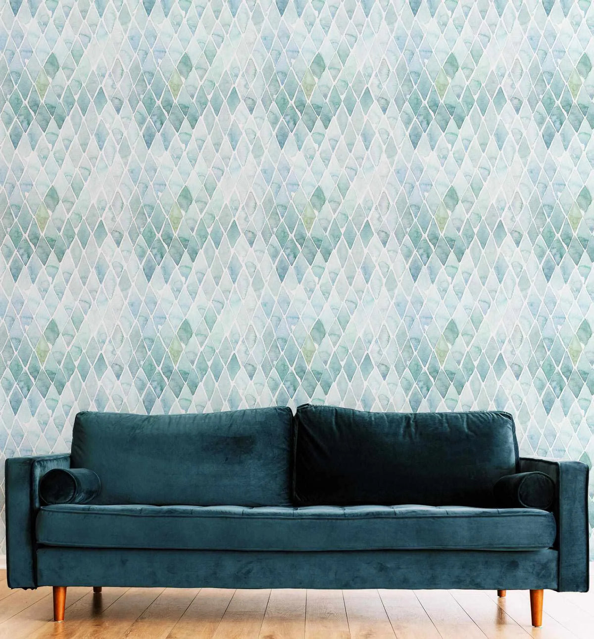 Seafoam Small Tile Wallpaper displayed on a wall behind a dark teal velvet sofa with wooden legs. The wallpaper features a repeating geometric pattern of small diamond shapes in varying shades of seafoam green, pale blue, soft gray, and cream, creating a subtle tonal variation across the surface. The tiles have a watercolor-like quality with soft, blended edges. The wood flooring and mid-century modern sofa styling emphasize the calming, muted color palette of the design.