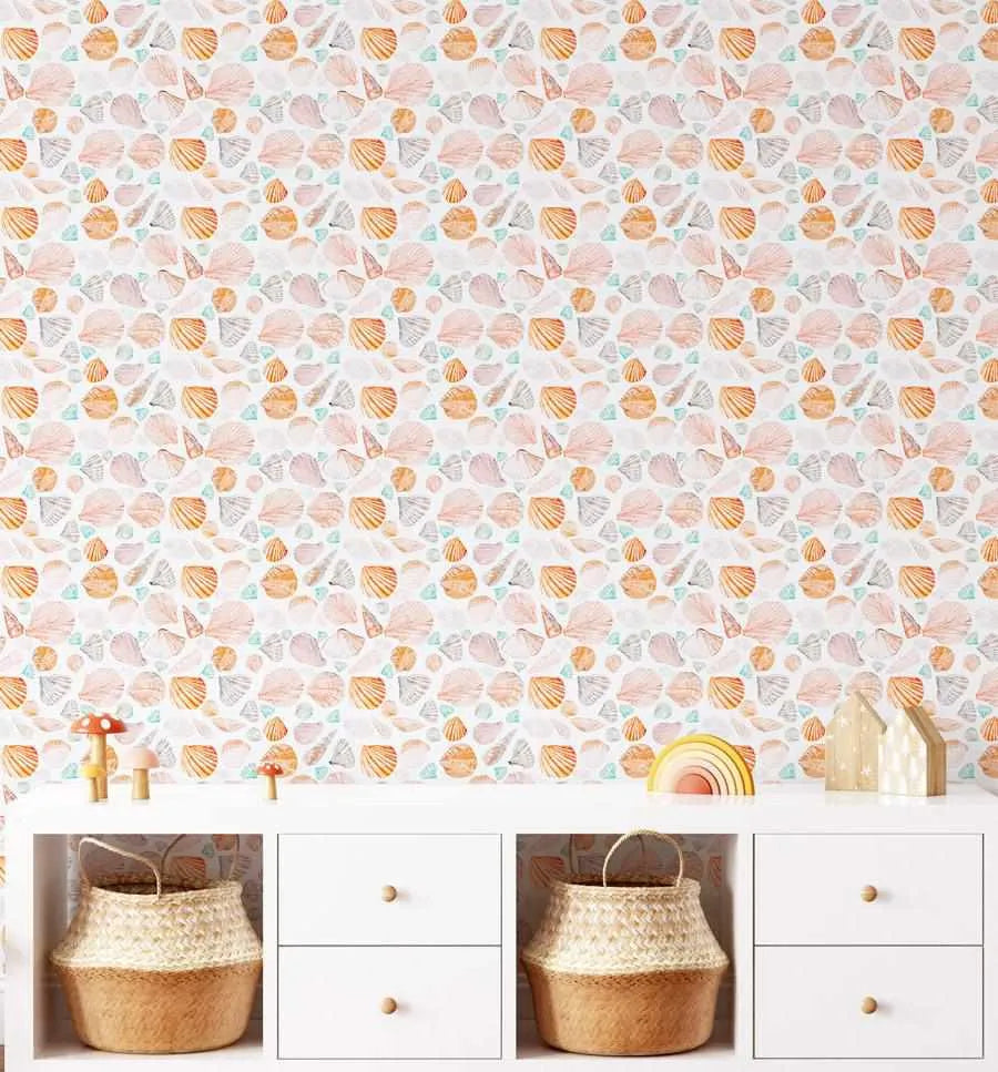 Lifestyle shot of a child's room featuring Seashells Wallpaper above a white storage unit. The wallpaper has a repeating pattern of various seashells in soft peach, muted orange, pale grey, and light teal, with a hand-painted, watercolour quality on a white background. The storage unit has two woven baskets and four white drawers with light wood knobs. On top of the unit are decorative wooden mushrooms, a rainbow toy, and two small wooden house ornaments.