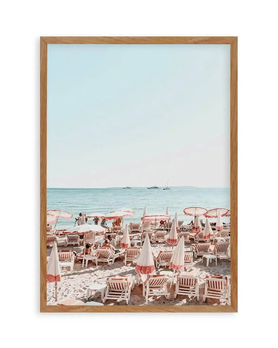 Framed photography art print of a vibrant beach scene with a natural wood frame and white border. The composition features rows of white and coral-striped beach chairs and closed umbrellas on light sand, with the turquoise ocean and distant boats under a pale blue sky.