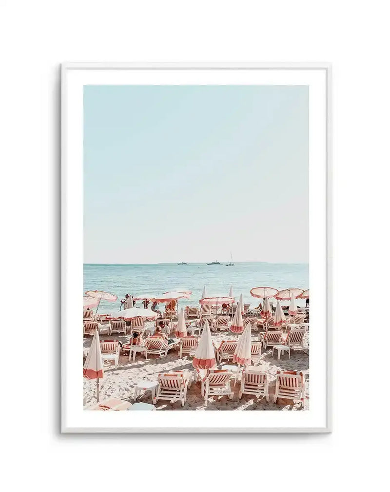 Framed photography art print of a vibrant beach scene with a white frame and white border. The composition features rows of white and pink striped beach umbrellas and lounge chairs on light sand, with the turquoise ocean and a clear pale blue sky in the background. Several boats are visible on the horizon.