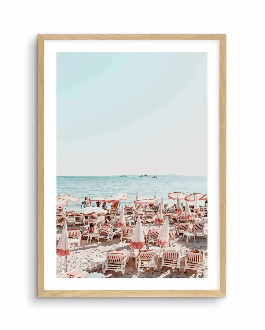 Framed photography art print of a vibrant beach scene with natural wood frame and white border. The composition features rows of white and pale pink striped beach umbrellas and lounge chairs on light sand, with people relaxing. Beyond the beach, the calm turquoise ocean stretches to the horizon under a clear, pale blue sky, with several boats visible in the distance.